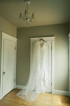 An empty room with light sage green walls, white doors, wood flooring, and sheer white curtains illuminated by a chandelier.