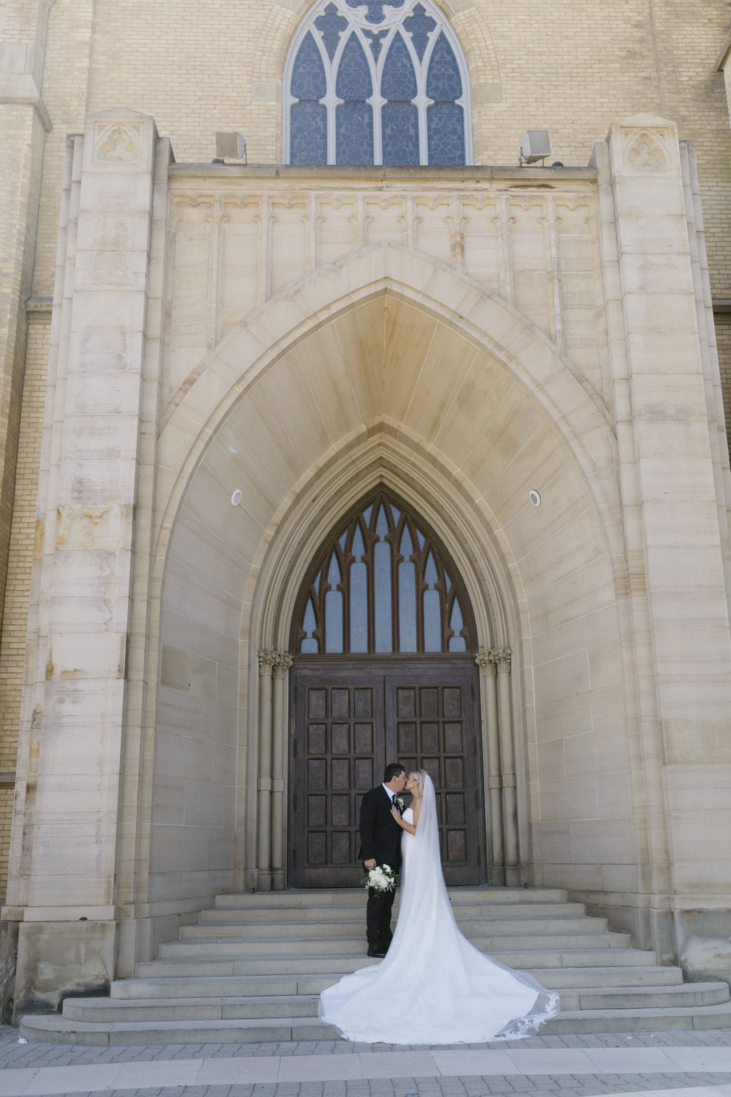 A wedding couple stands in front of a grand gothic church archway with ornate stone details and wooden doors.
