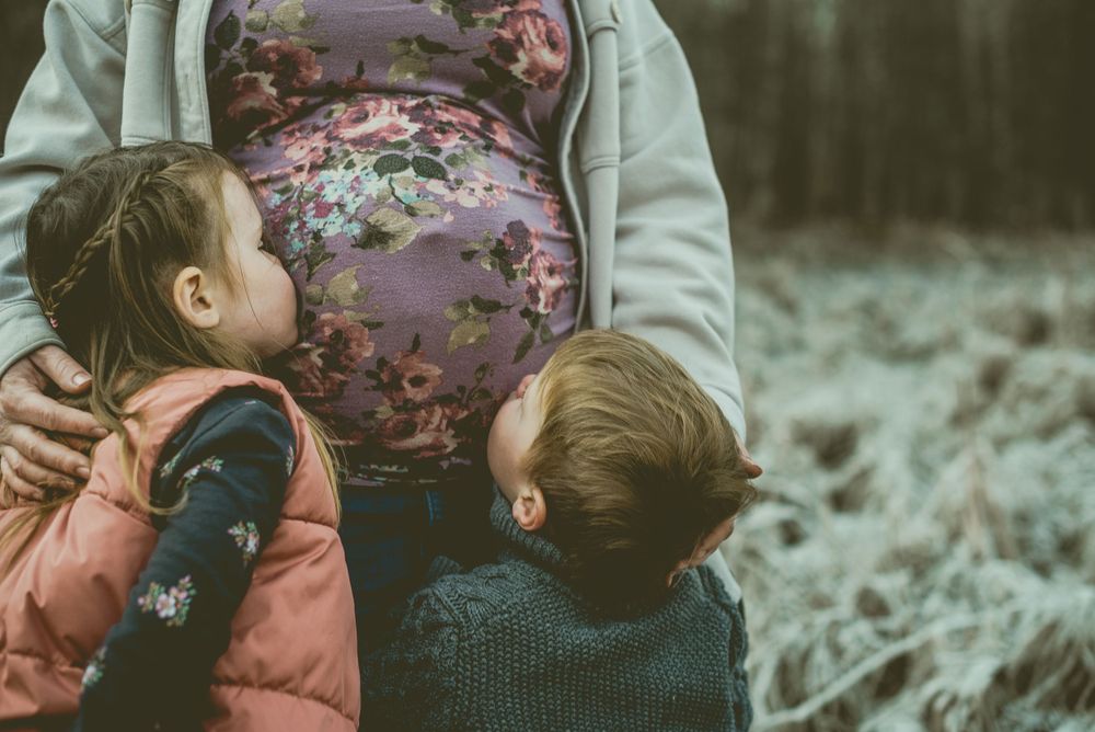 Children in winter coats embrace a floral-dressed pregnant belly while standing outdoors on frosty ground.