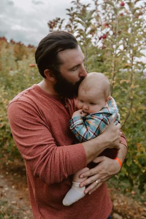A tender moment captured at an orchard as wearing a coral sweater gently embraces and kisses an infant in plaid. Fatherhood, Father and son