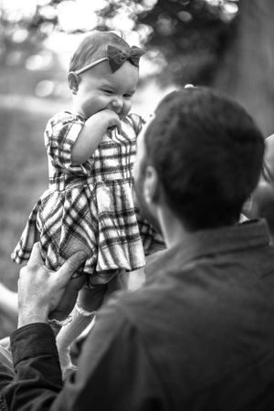 Black and white photo of a small child in a plaid dress and bow being held up in the air during a playful moment. Fatherhood, Family