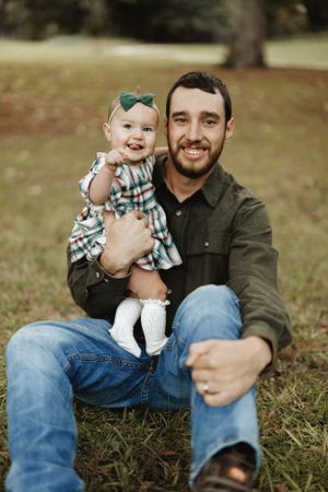 A heartwarming outdoor portrait shows a baby in plaid dress with a green bow sitting on an adult's lap in a grassy setting. Fatherhood