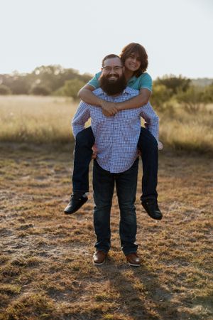 A couple enjoying a playful piggyback ride in a sunlit field during golden hour. Father and son, family, fatherhood