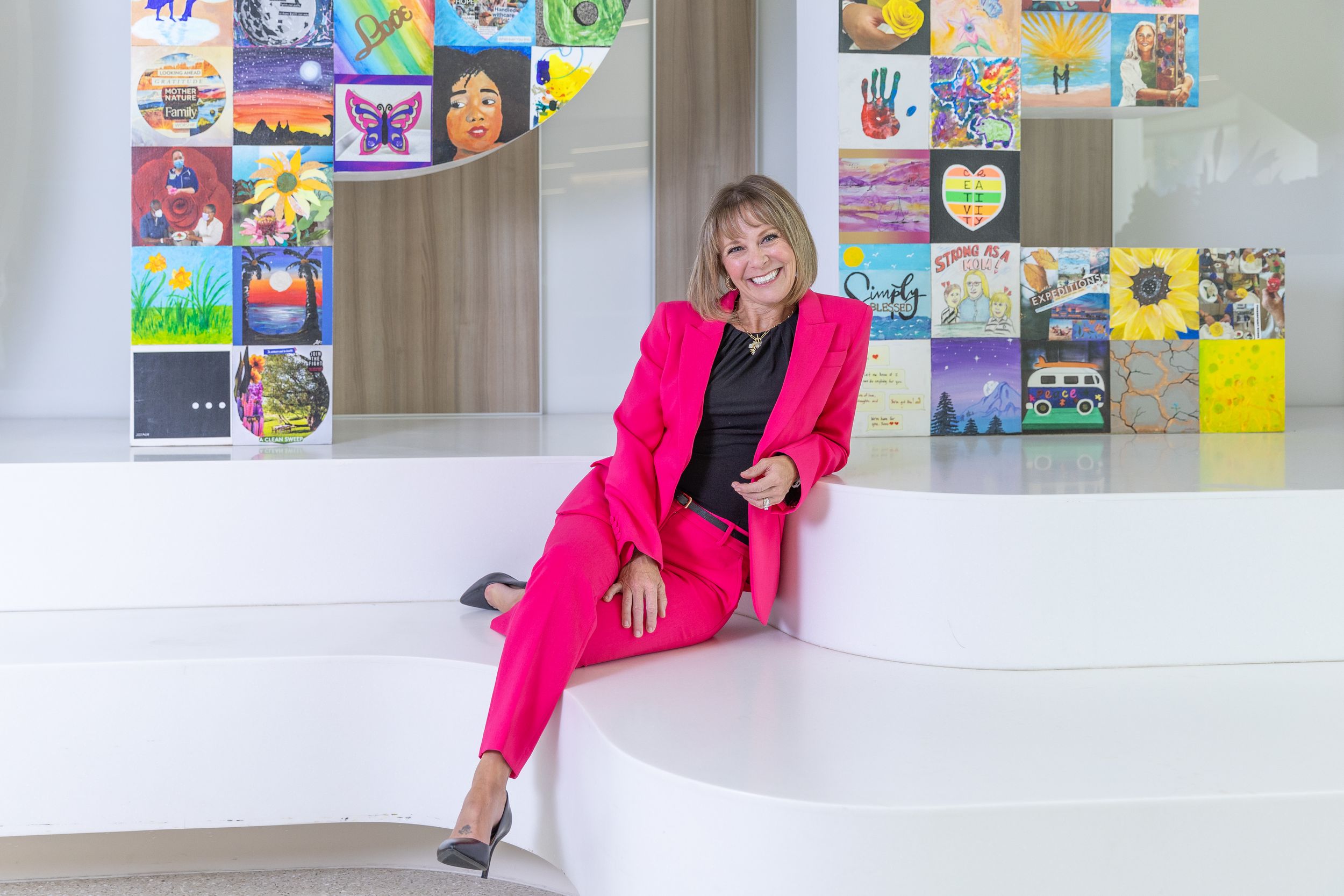 A person in a bright pink blazer sits on a white modern bench against a wall decorated with colorful posters and artwork.