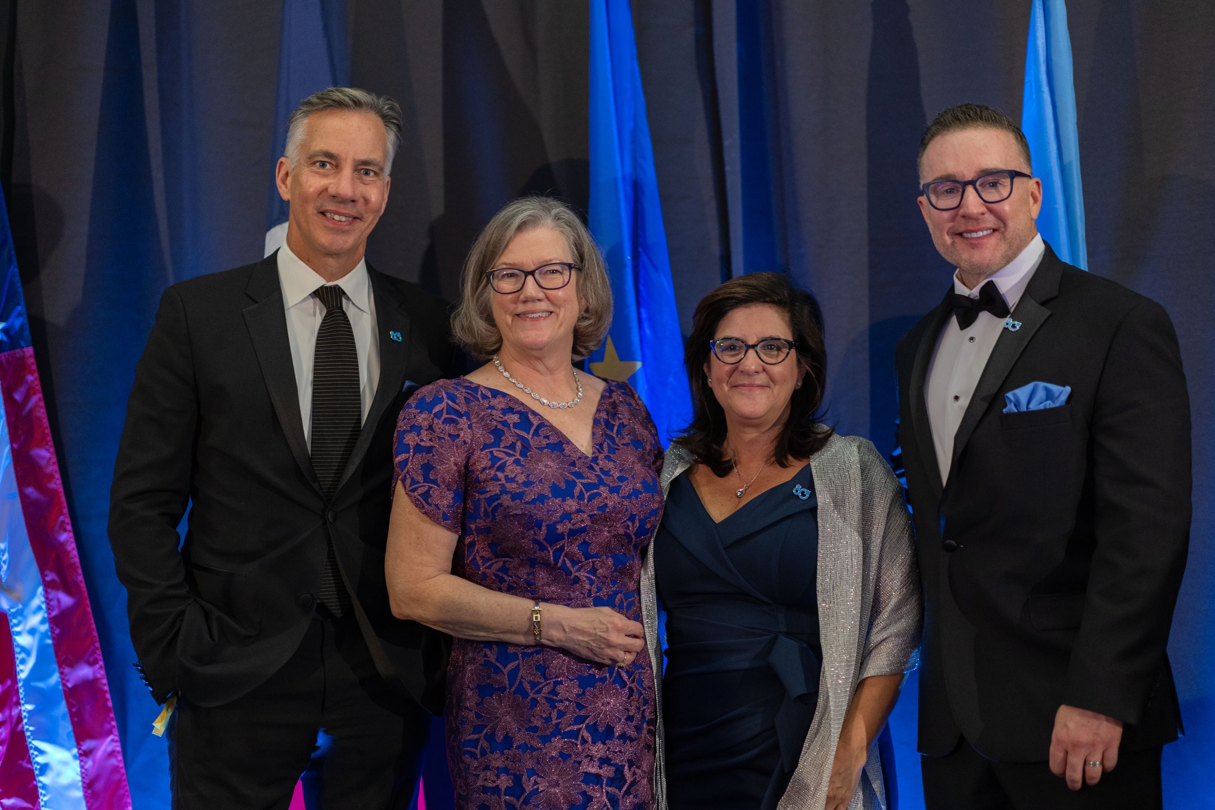 A group of formally dressed people stand together at an event with blue lighting in the background.