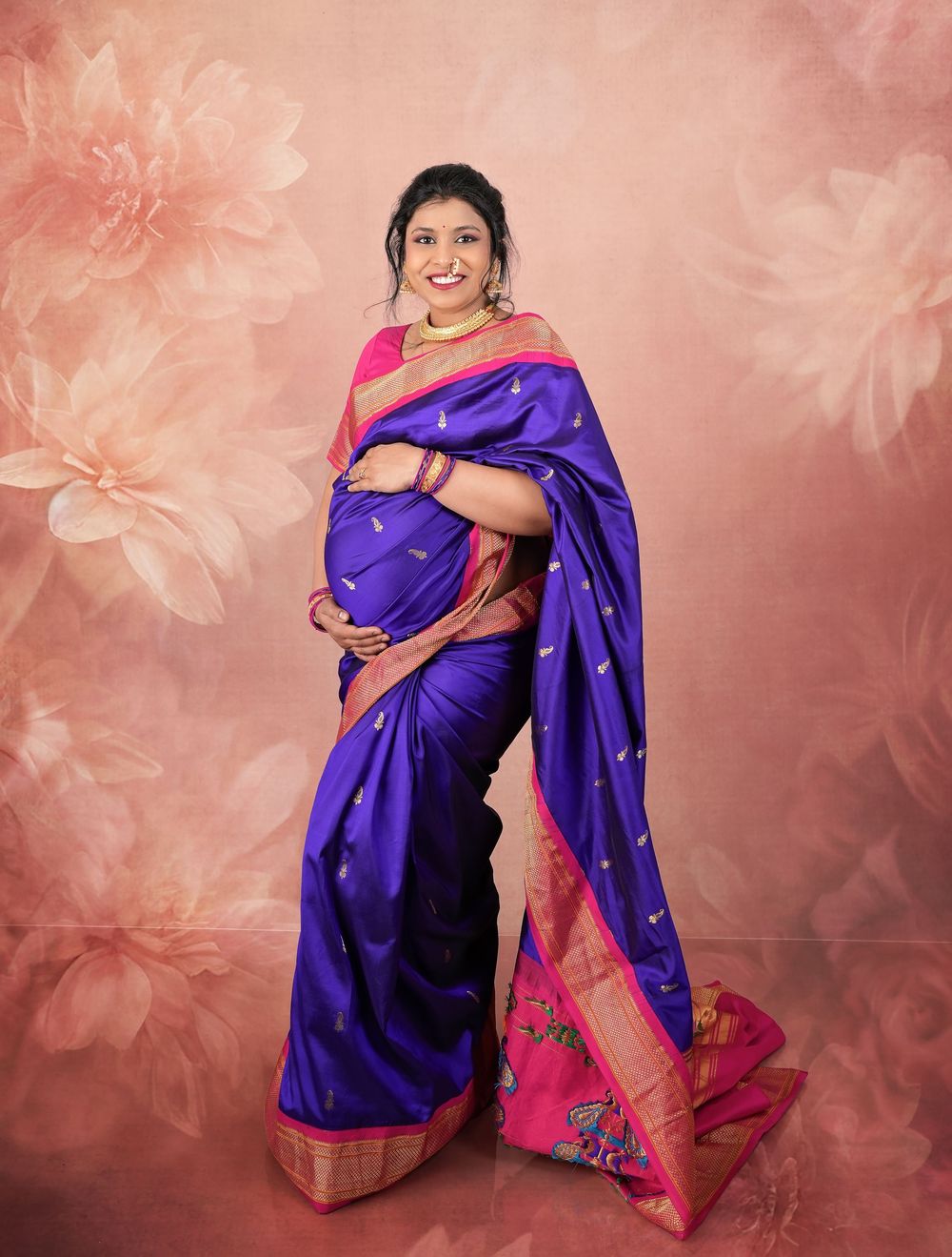 Expectant mother in royal blue silk saree with pink border poses against floral background.