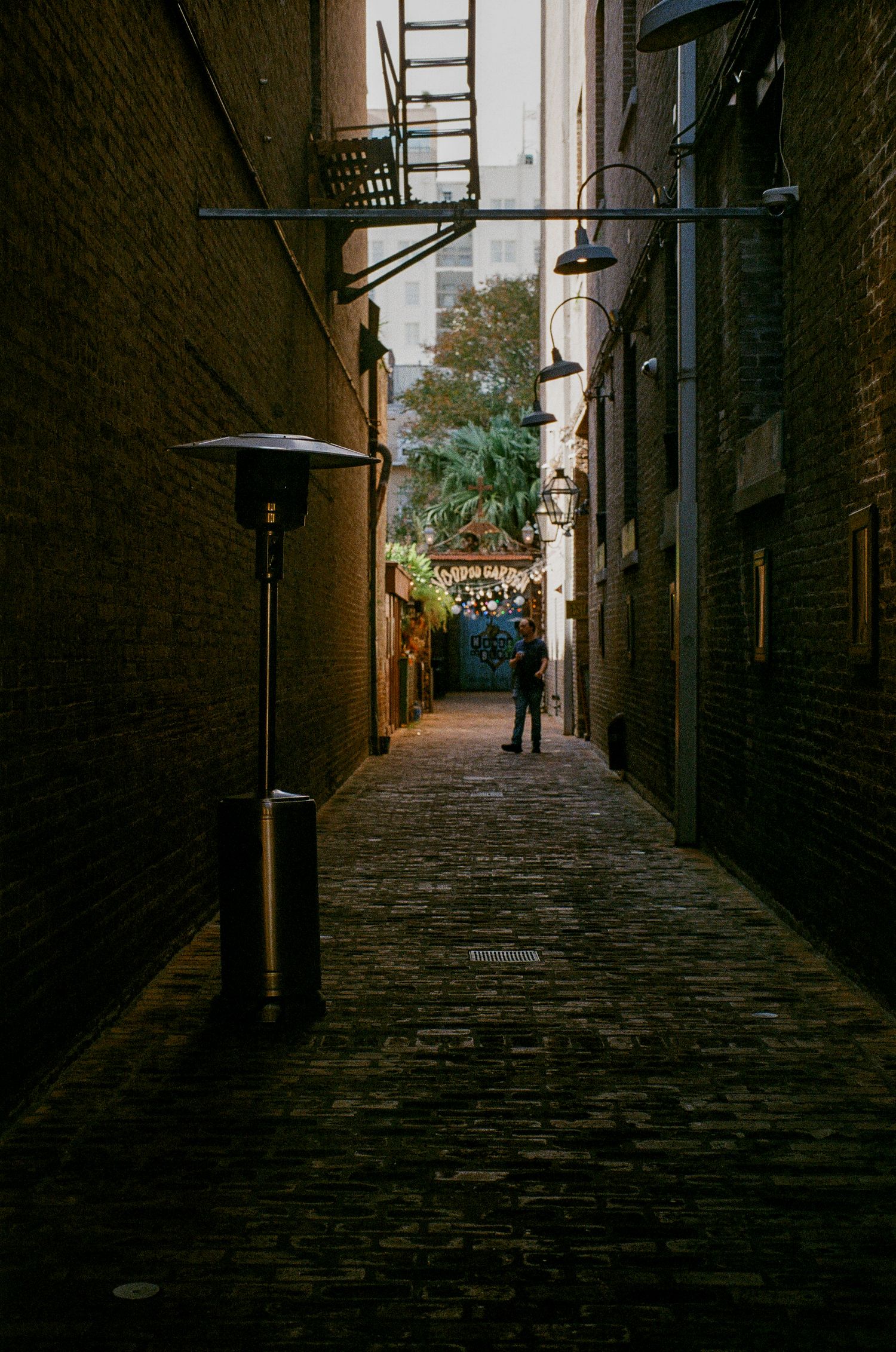 A narrow brick alleyway with string lights leads to a distant figure walking beneath industrial lamps.