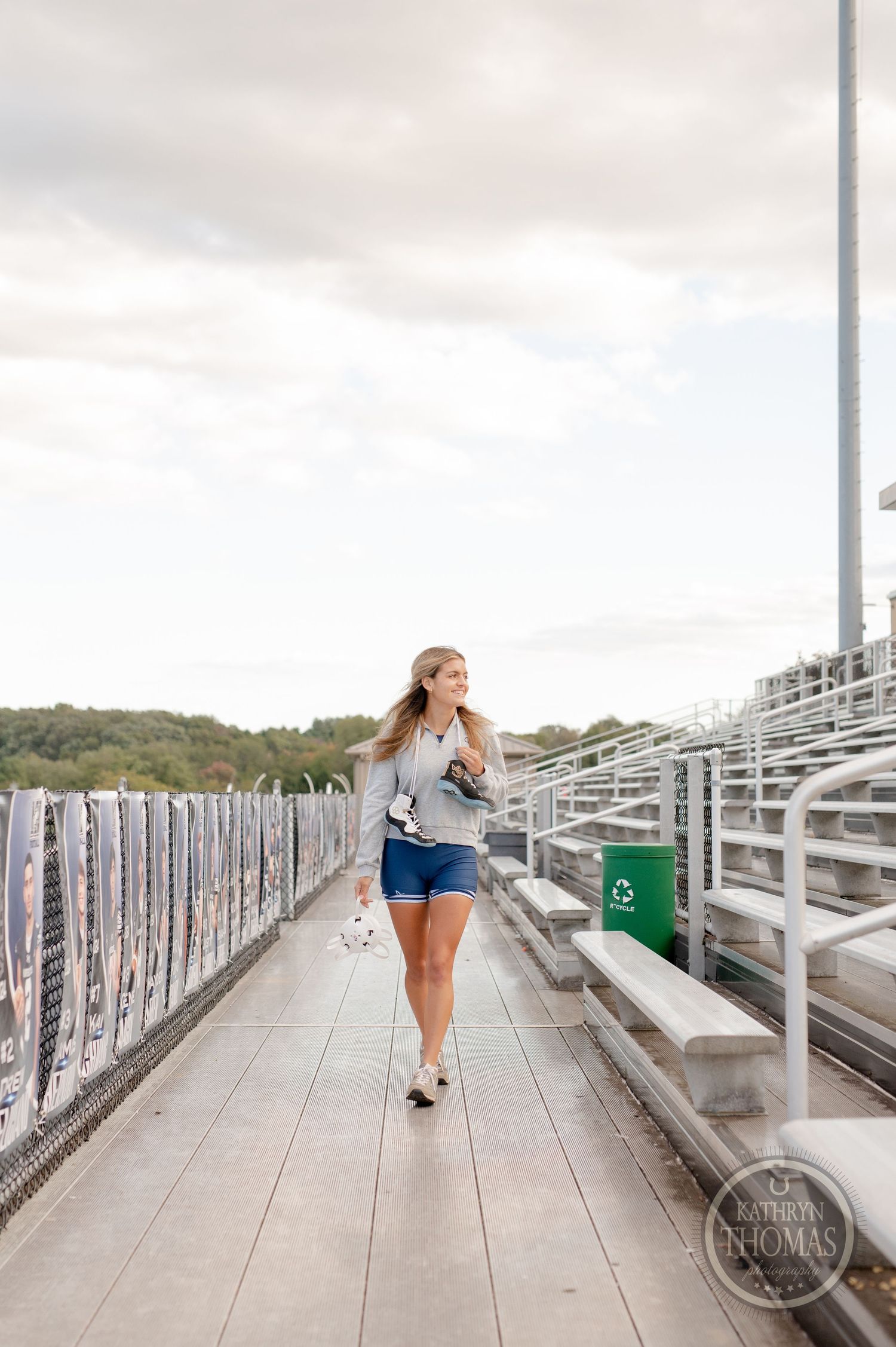 Athlete in grey sweatshirt and blue shorts walking along stadium bleachers holding sports equipment.