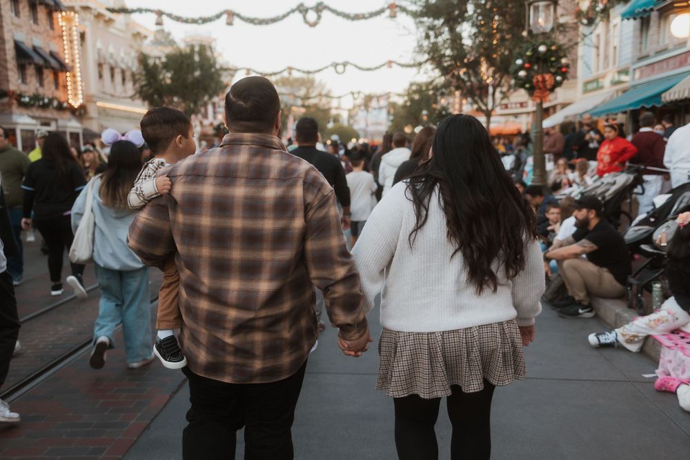 Two people hold hands while walking through a crowded Disney Main Street decorated with overhead lights and festive decor.