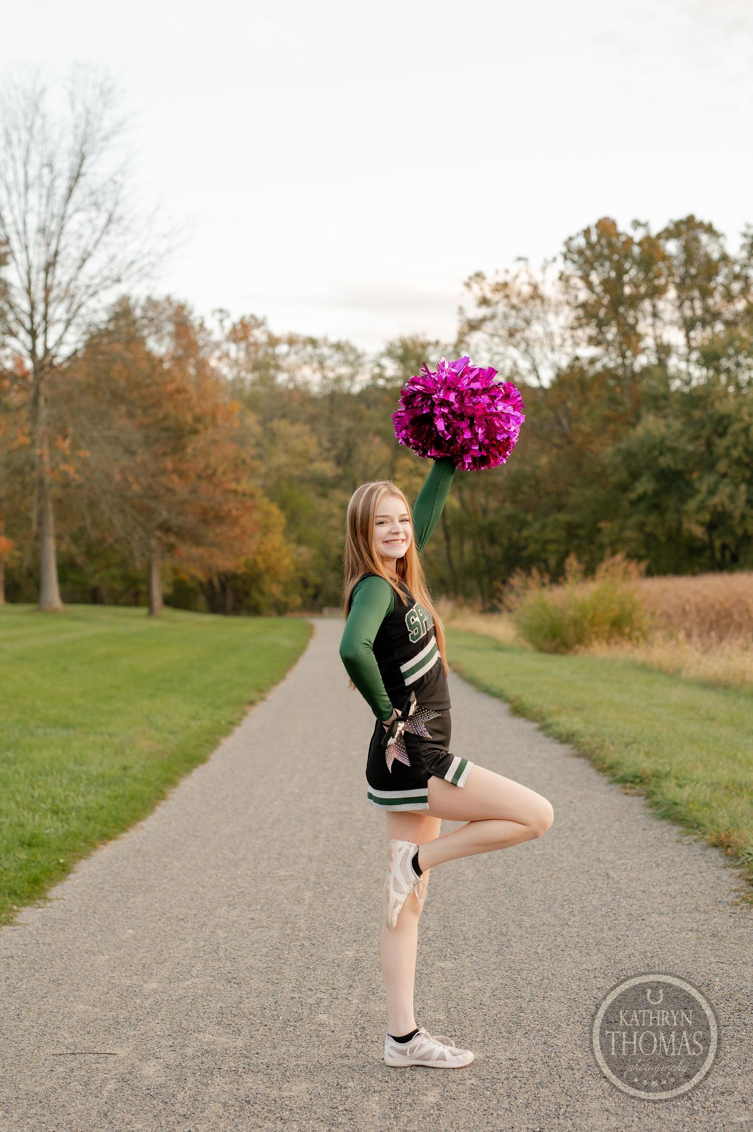 Cheerleader poses with bright pink pom pom on path surrounded by fall foliage.