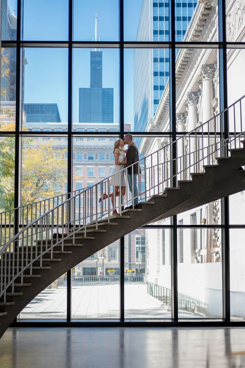 Couple stands on dramatic curved staircase against Chicago skyline viewed through large window panels.