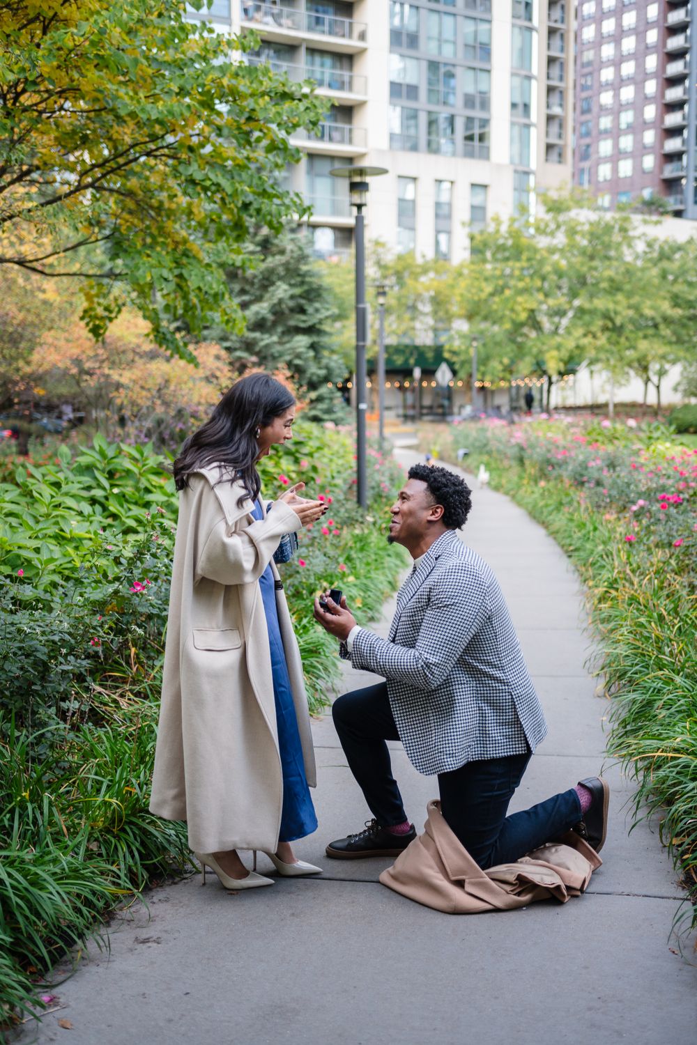 Marriage proposal scene in lush garden path with city buildings in background.