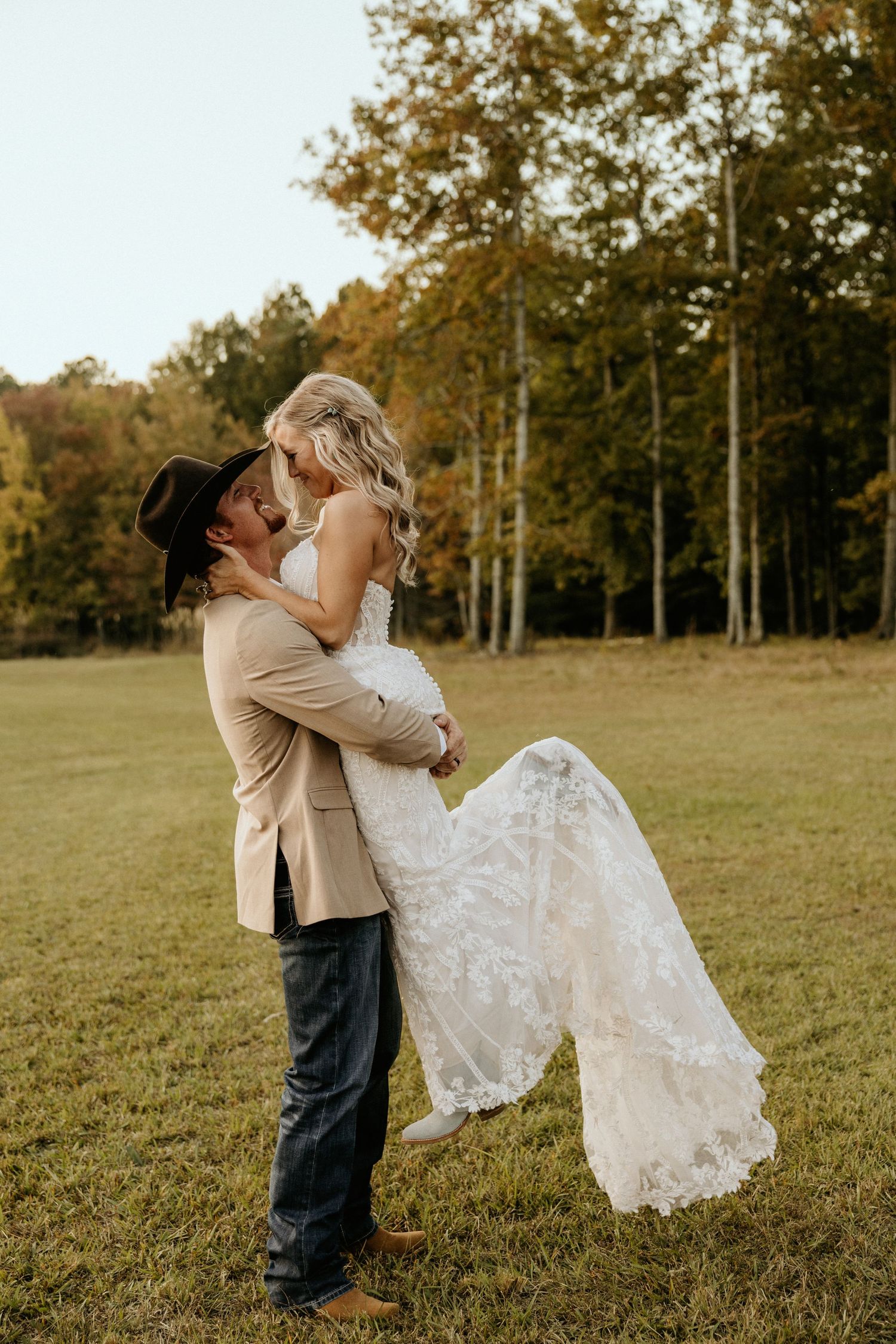 Romantic wedding photo with groom lifting bride in lace dress during outdoor countryside photoshoot.