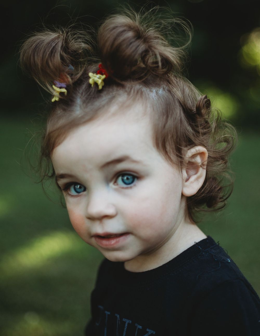 Portrait of toddler with curly brown hair and blue eyes wearing a dark top outdoors with green foliage in background.