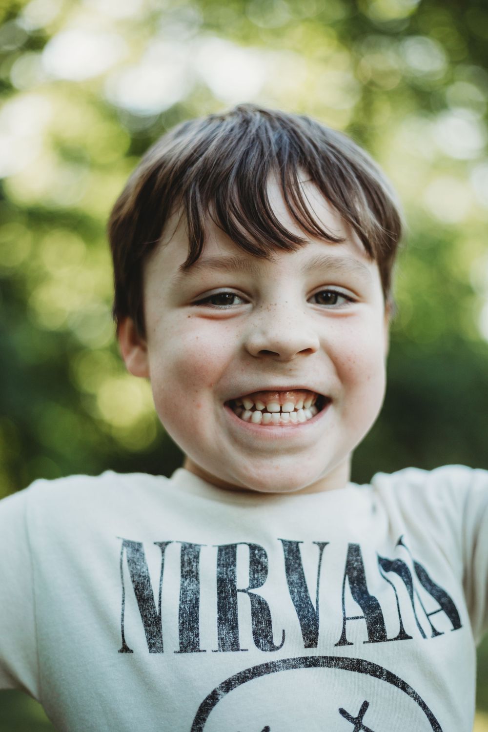 Close-up portrait of a smiling child wearing a Nirvana band t-shirt outdoors with bokeh background.