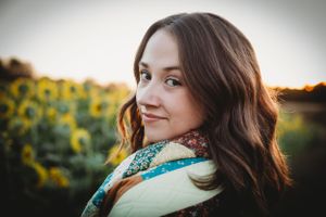 Portrait photo of a smiling woman with dark wavy hair against a background of sunflowers at sunset.