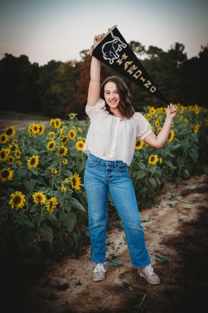 Woman in white eyelet top and jeans posed joyfully in a sunflower field holding a graduation cap.