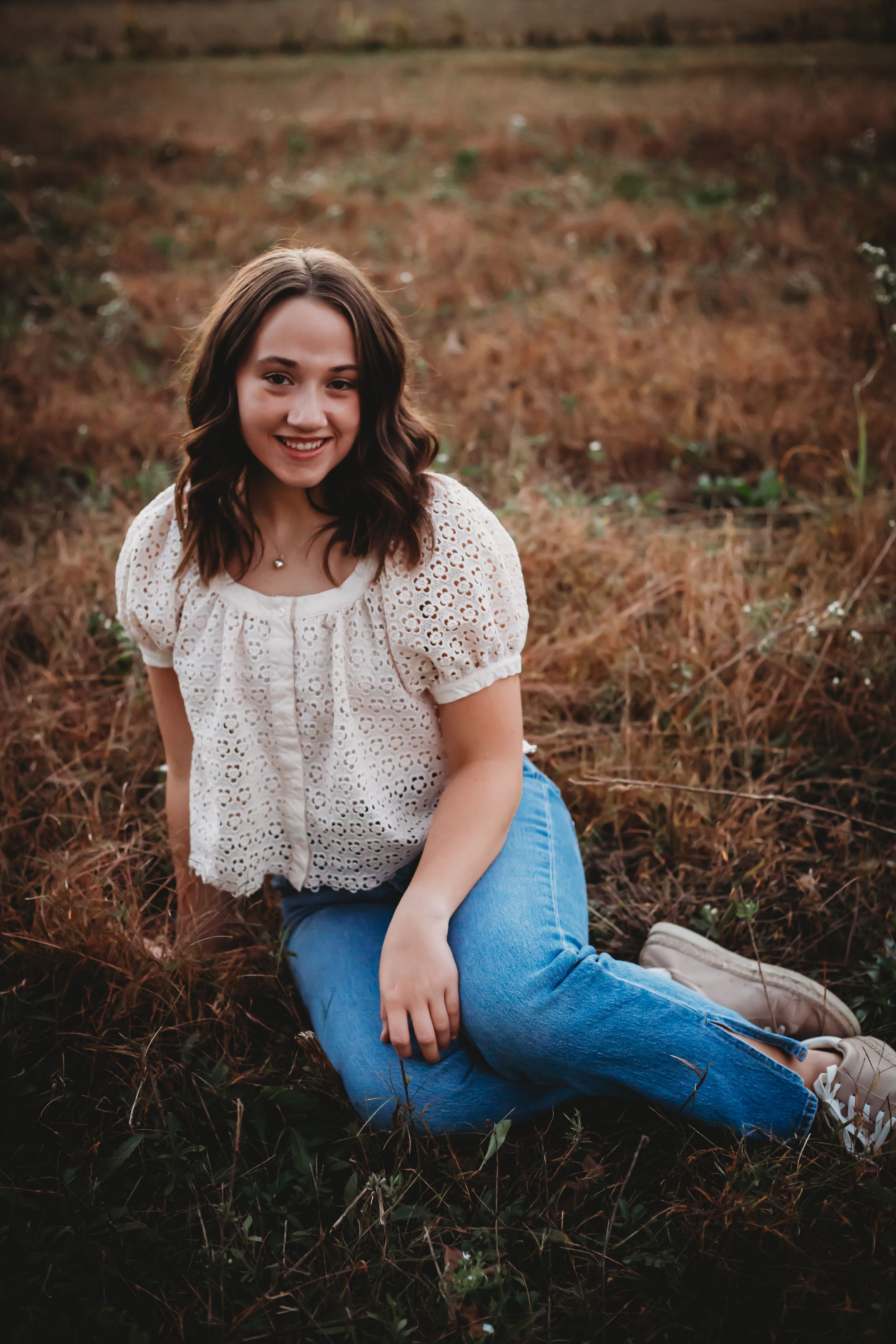 Woman wearing white eyelet top and jeans sitting on rustic ground during golden hour photoshoot.