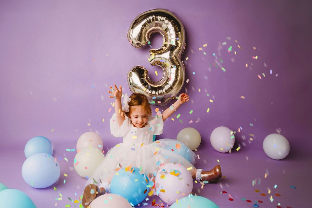 Birthday party setup with metallic number balloons, pastel decorations, and confetti against a purple backdrop.