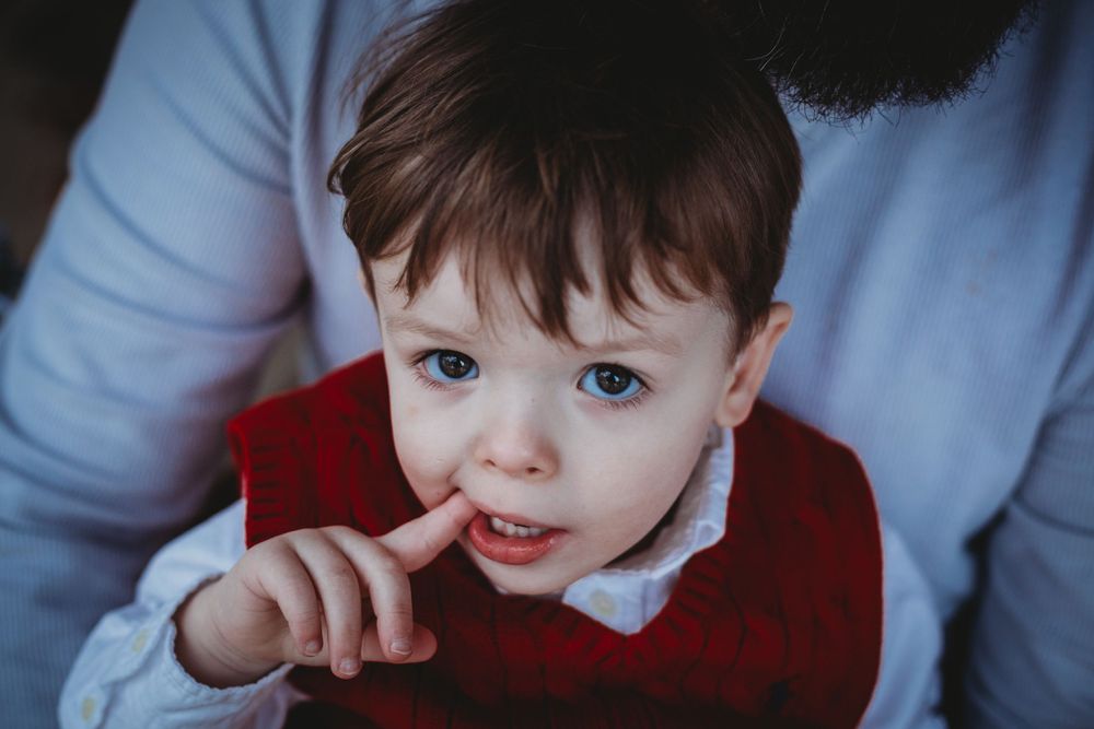 Toddler in a red sweater with finger near mouth smiling at the camera.