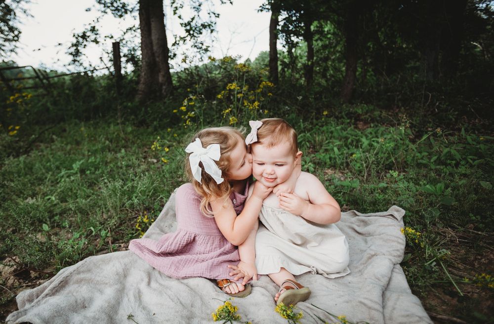 Two small children in dresses sit close together on a blanket outdoors in a wooded setting.