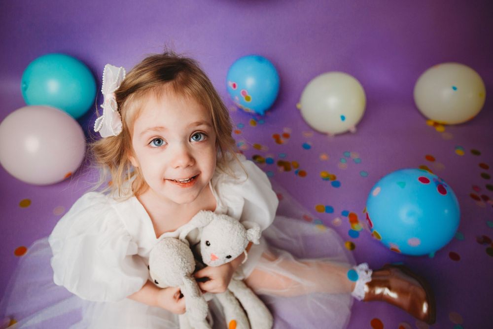 A child in a white dress poses with a teddy bear surrounded by blue and white balloons and confetti.