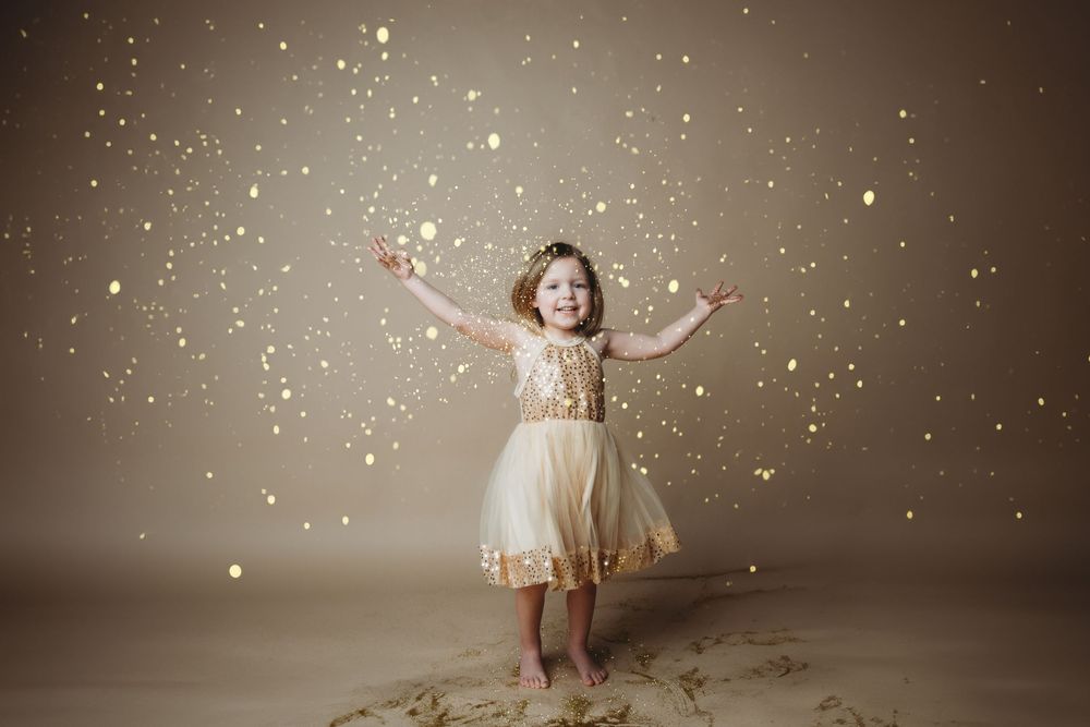 A whimsical photo series shows a child in a white dress playing with falling glitter and sparkles against a brown backdrop.