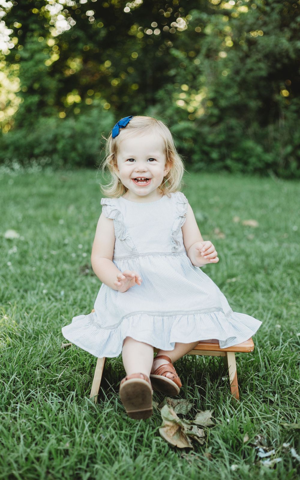 Young child in white dress sitting on wooden stool outdoors with bright natural background.