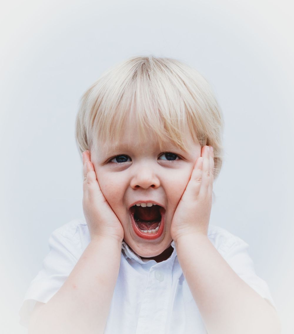 A child with hands on cheeks making an excited, open-mouthed expression against a light background.
