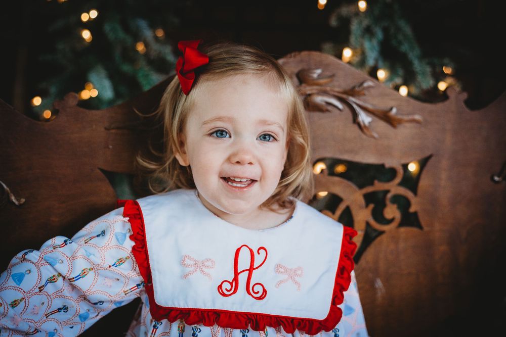 Child wearing holiday dress with monogrammed bib smiling with Christmas lights in background.