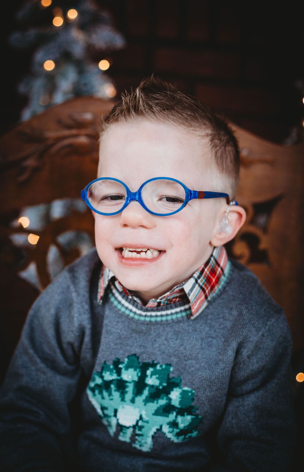 Smiling child in blue glasses and gray sweater with holiday lights in background.