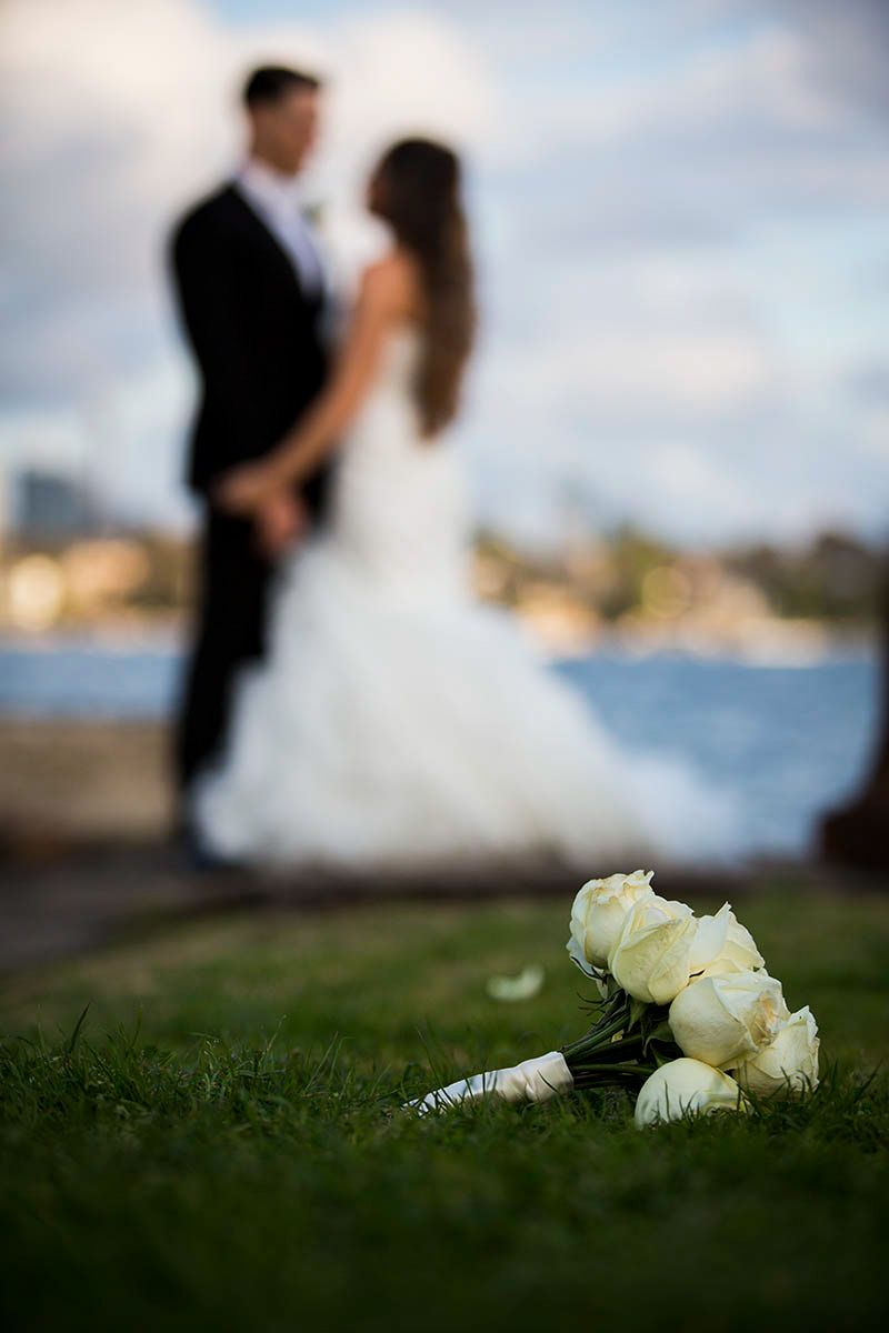 A white bridal bouquet lies on grass with a blurred waterfront scene in the background.