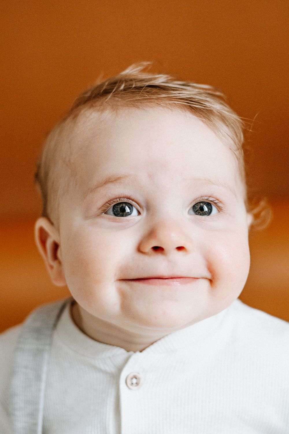 Close up portrait of a happy baby with big eyes wearing a white button-up shirt against an orange background.