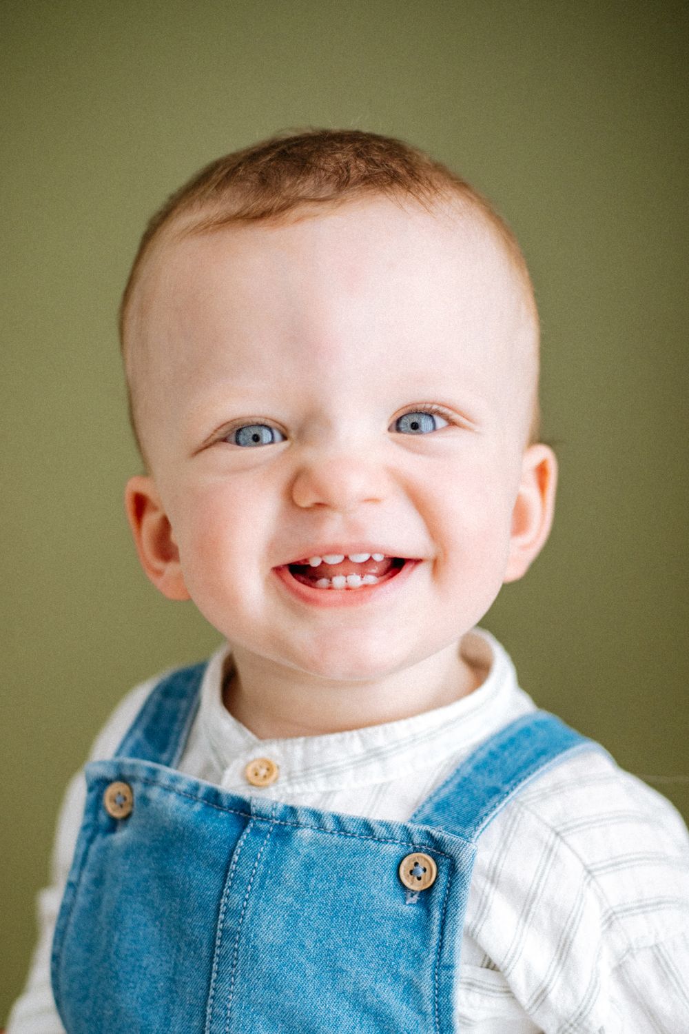An adorable baby with bright blue eyes and a big smile wearing denim overalls and a white shirt against a green background.