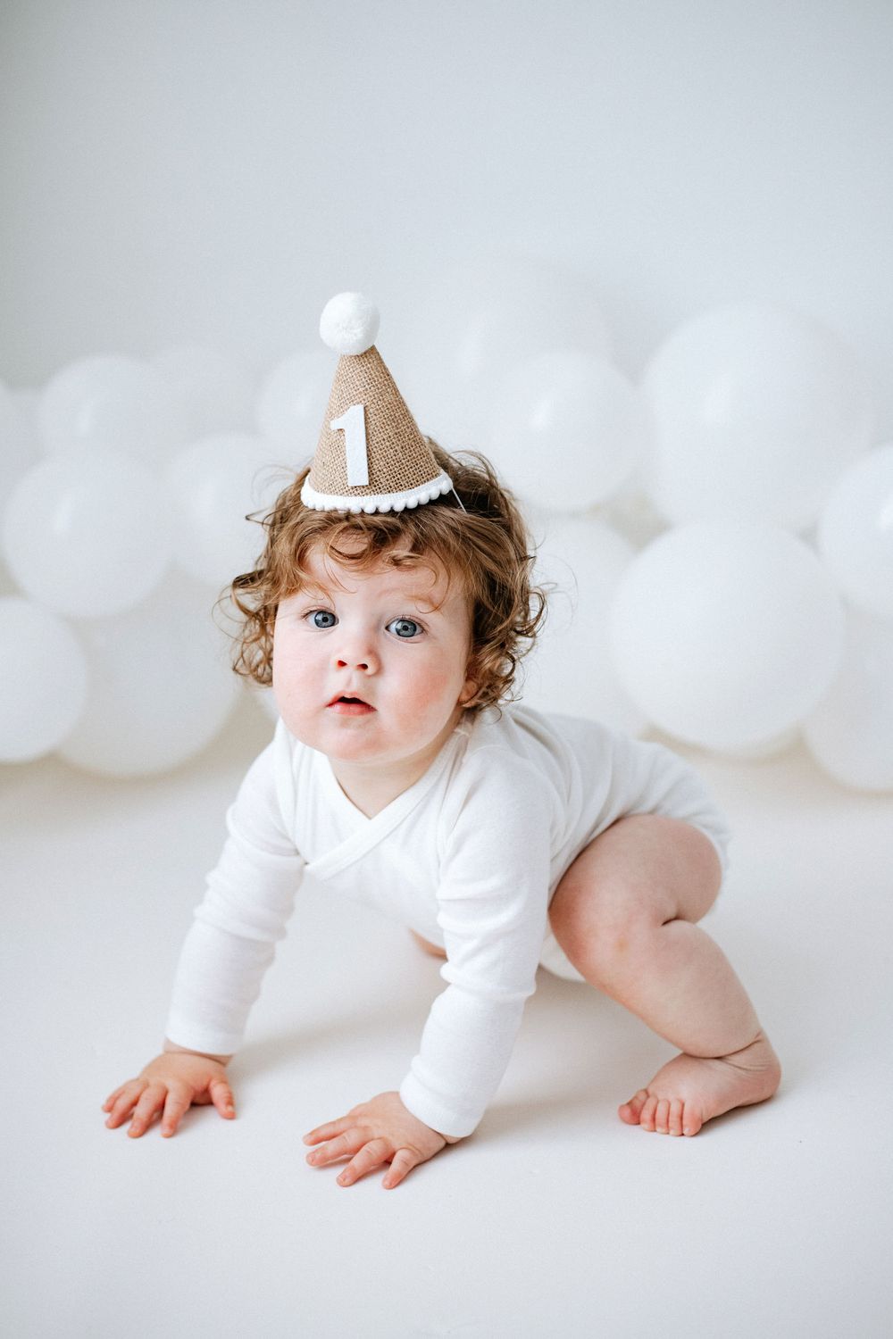 Small child in white bodysuit wearing a beige first birthday hat crawls on white backdrop with balloon decorations.