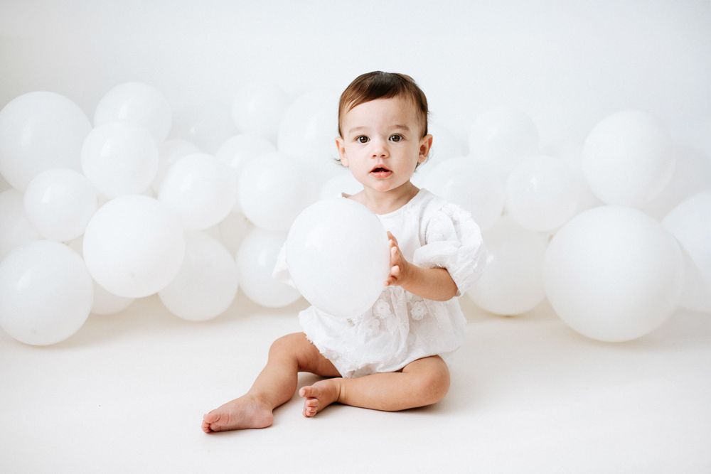 Baby wearing white bodysuit sits on white floor with balloon decorations in background.