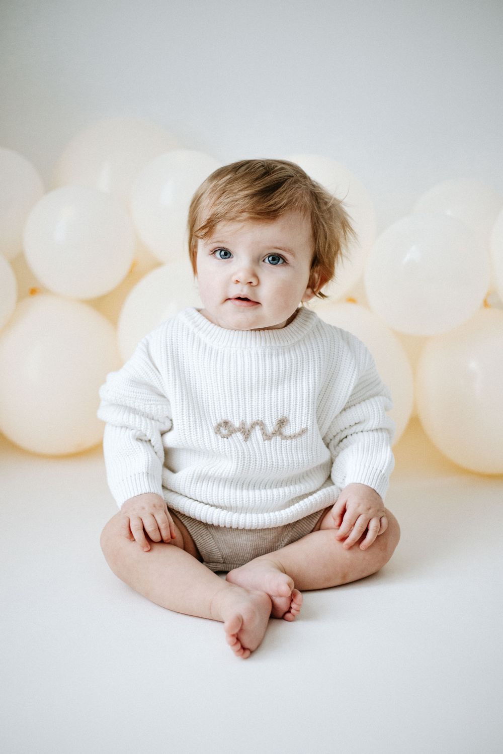 Small baby wearing white knit sweater with 'one' text sitting on floor with white balloons in background.