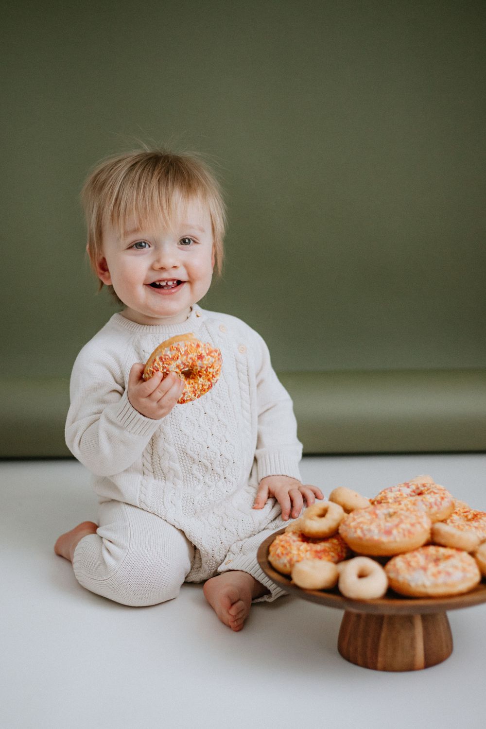 Small child in white outfit sits beside wooden cake stand filled with pink frosted donuts on white surface.