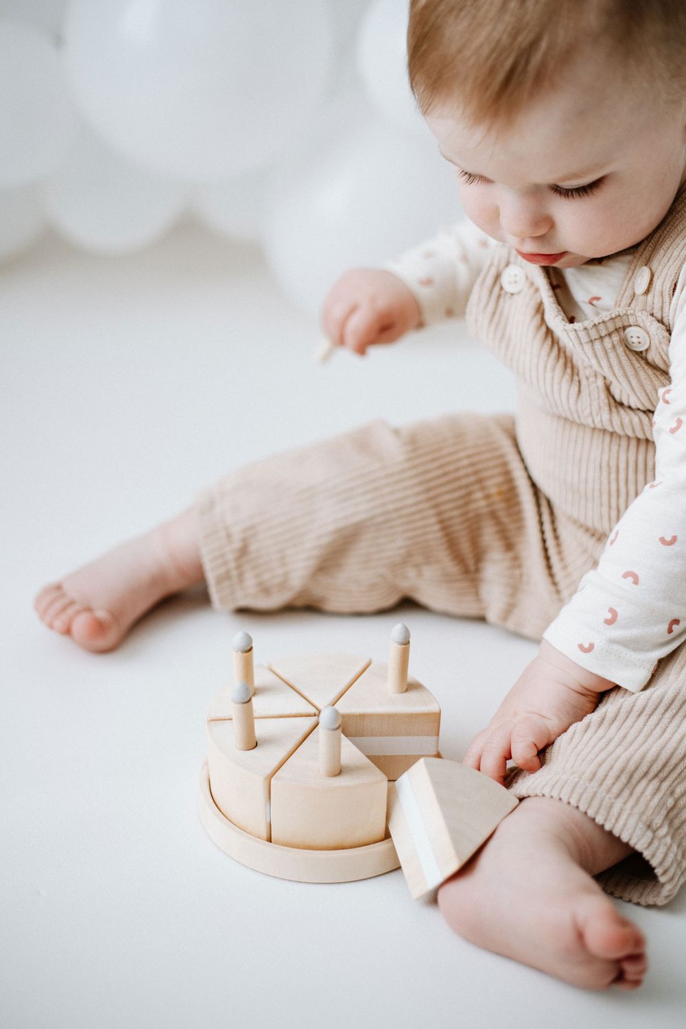 Small child in beige ribbed outfit playing with a wooden circular stacking cake toy on white background.
