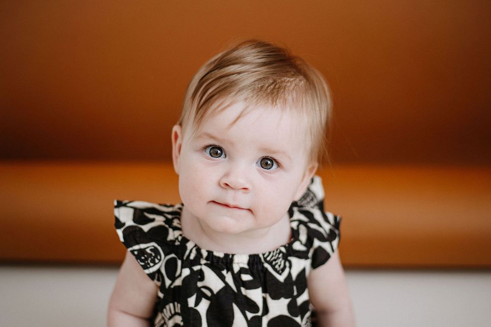 A young baby in a black and white floral dress poses against a warm brown background.