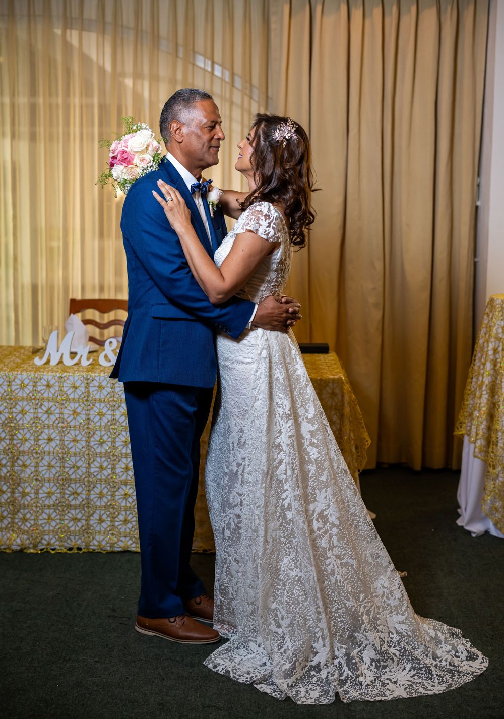 Couple in wedding attire sharing romantic moment against beige curtain backdrop.