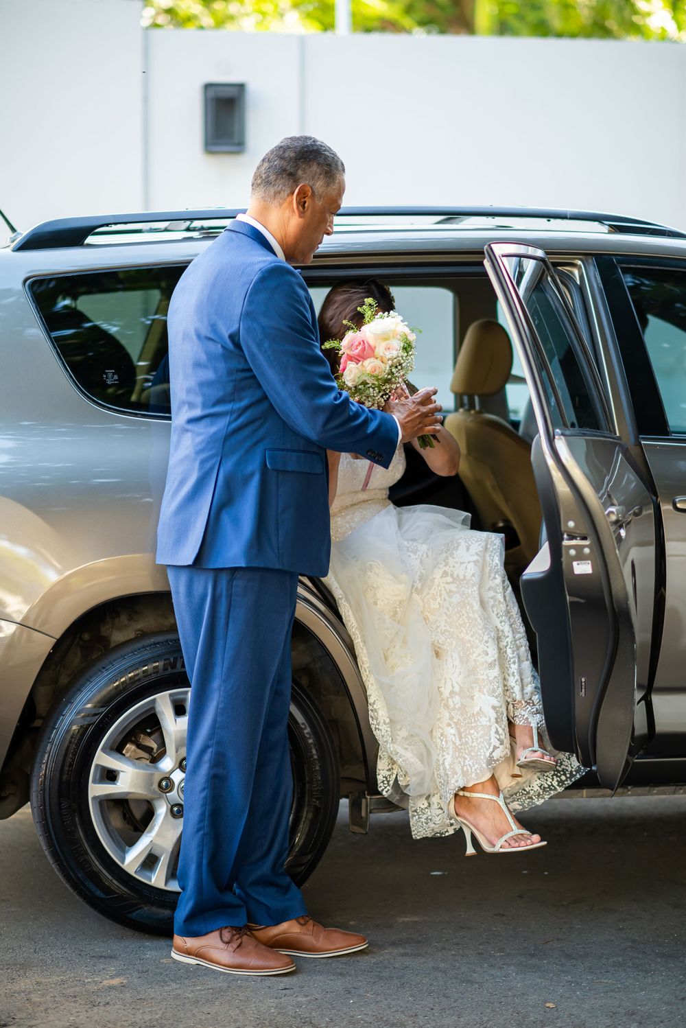 Groom in blue suit helping bride into car after wedding ceremony.