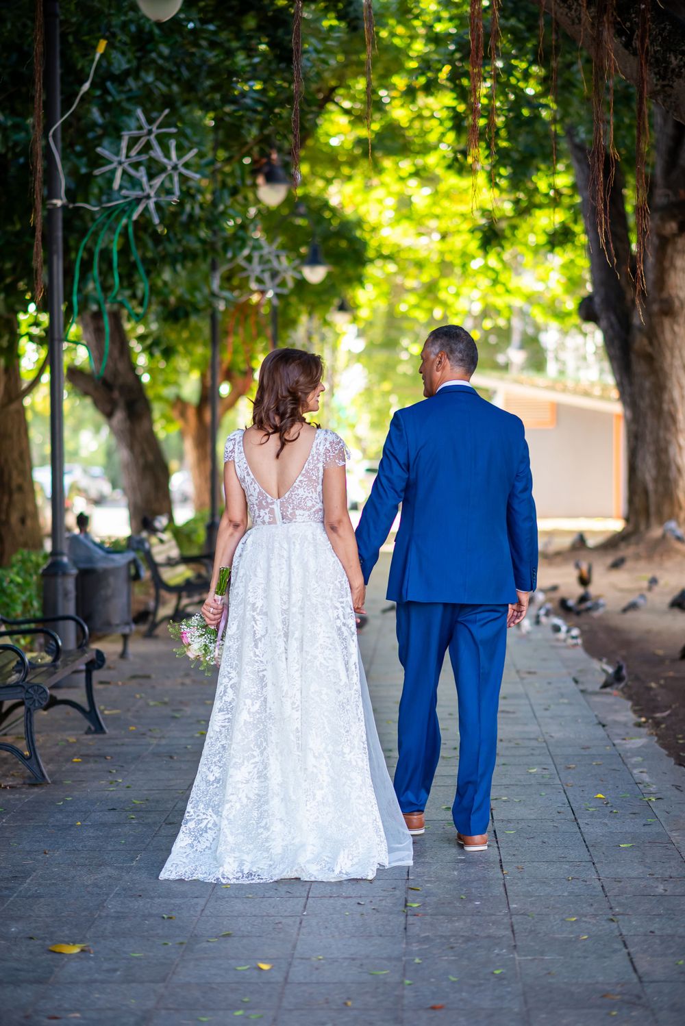 Newlywed couple walking down tree-lined path, bride in lace gown and groom in blue suit viewed from behind.