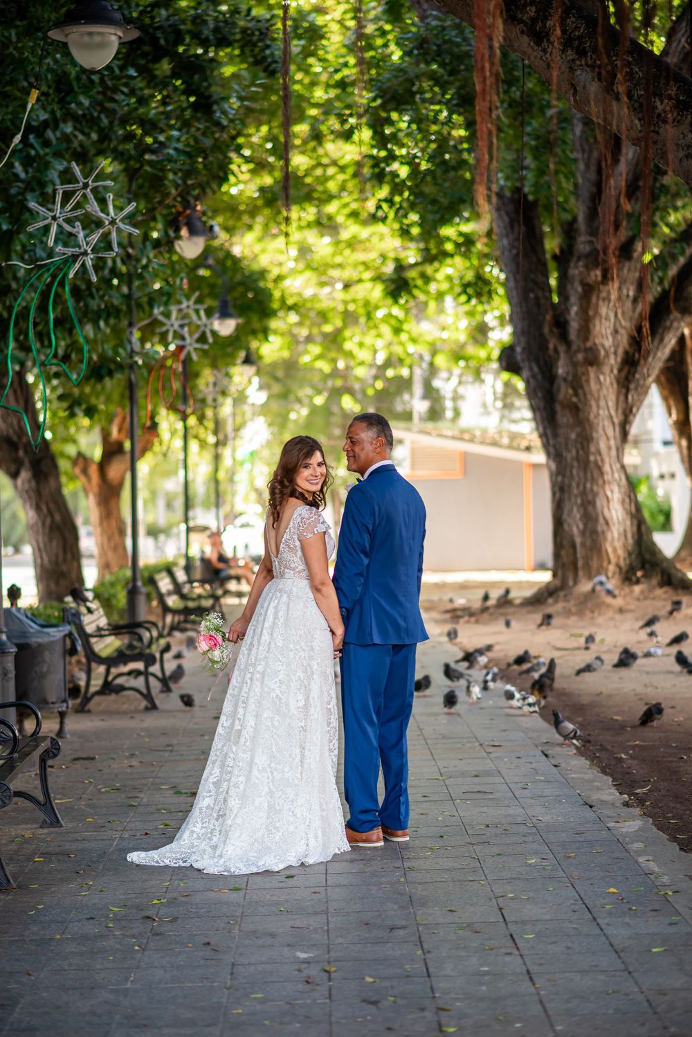 Bride and groom share romantic moment on tree-lined walkway with park benches.