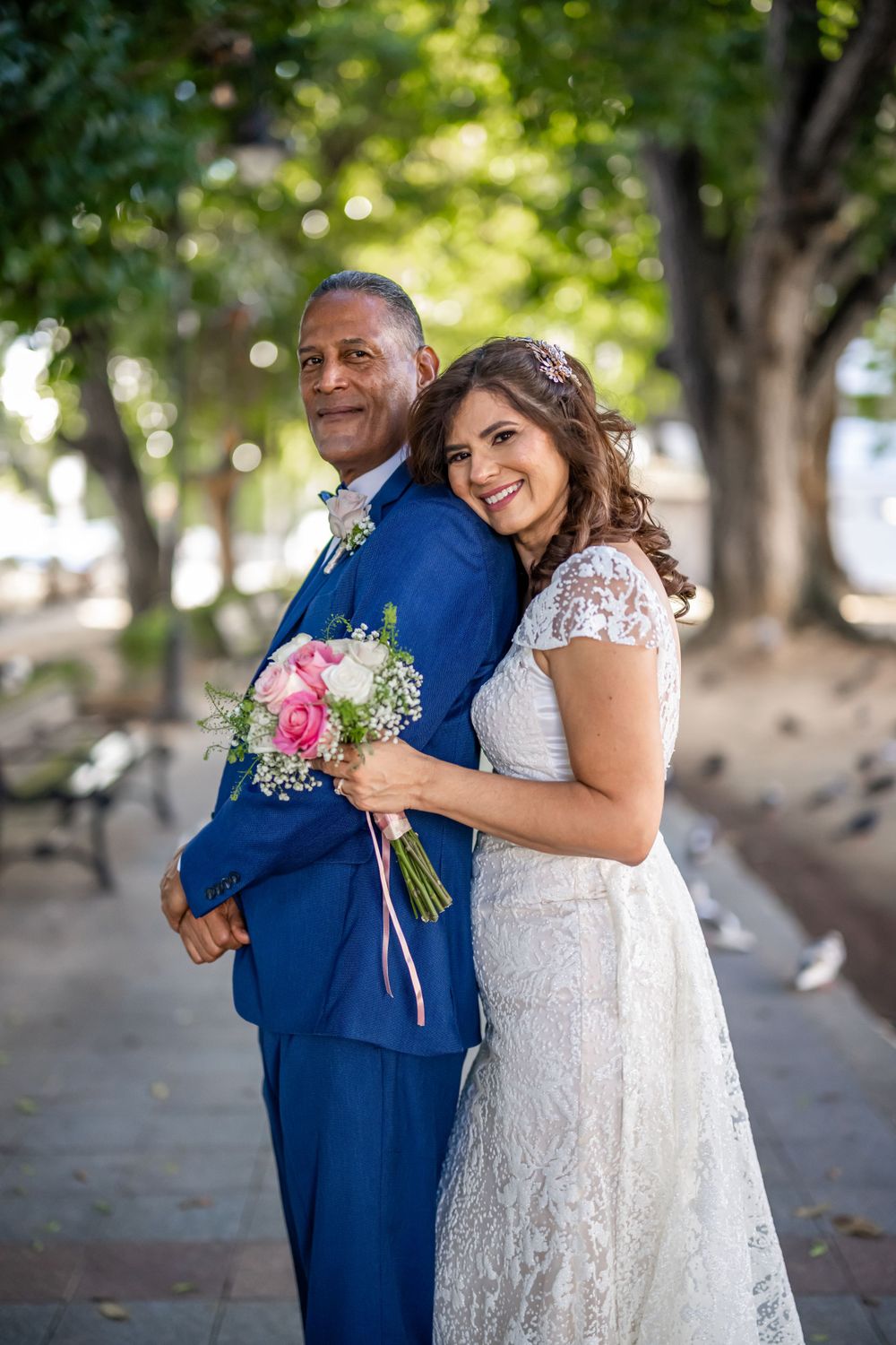 Happy couple poses in park, bride holding bouquet while leaning against groom in blue suit.