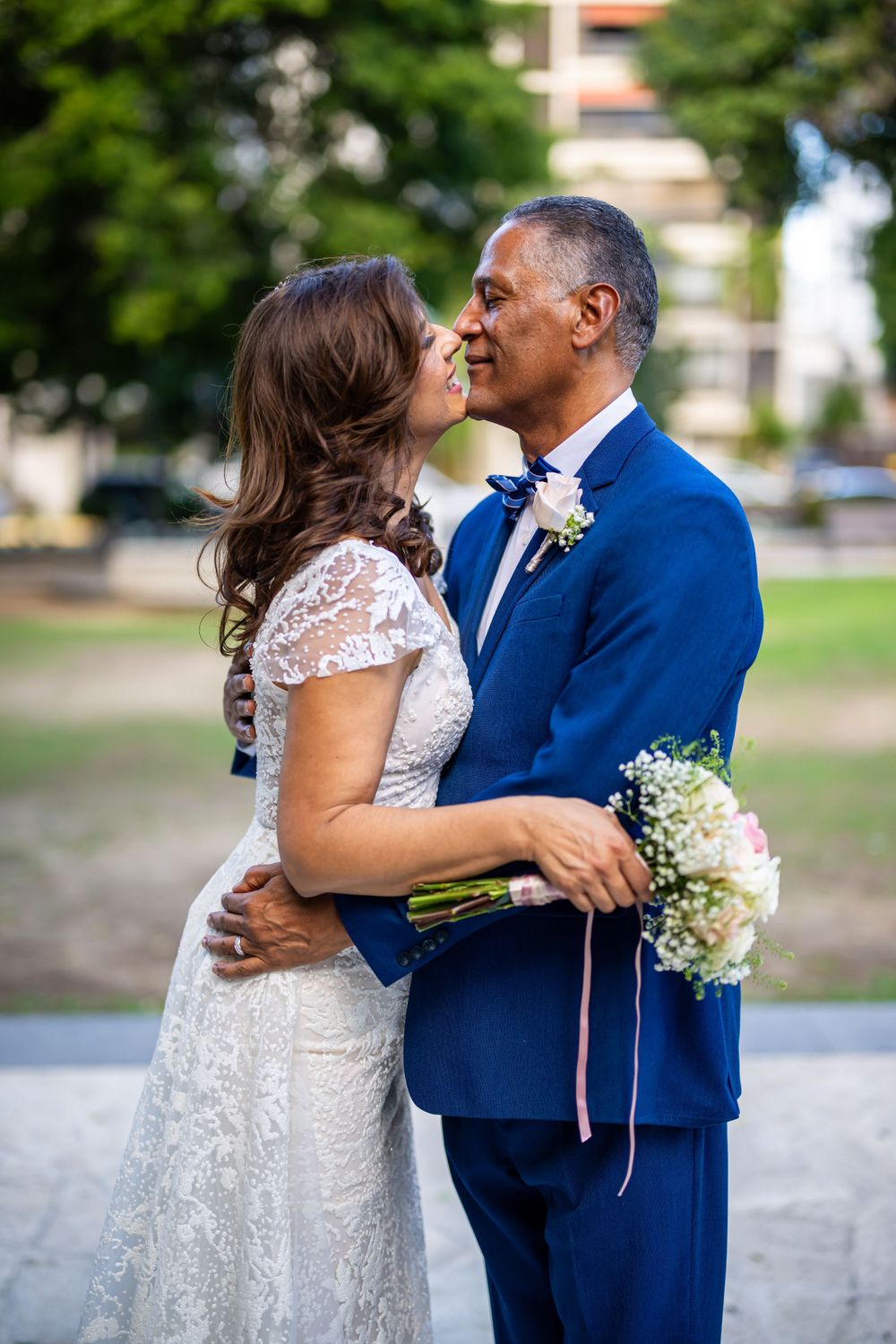 Intimate moment between newlyweds as they embrace in park with urban backdrop.