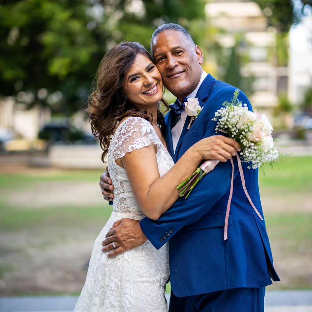 A romantic wedding couple embraces outdoors, with the bride wearing a lace dress and groom in navy blue suit.