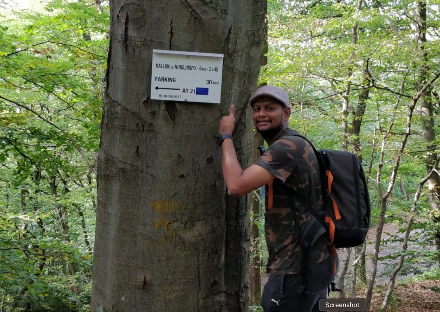 A hiker with a backpack points to a trail marker sign mounted on a tree in a lush green forest.