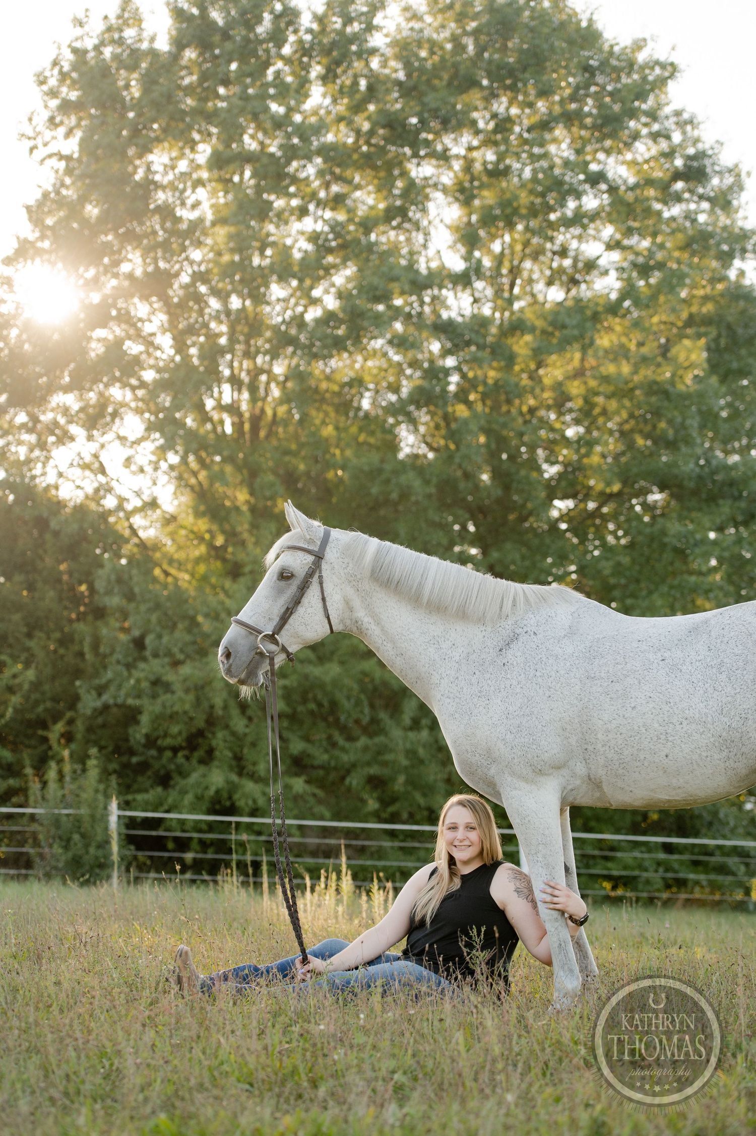 A person sits in a grassy field beside a white horse during sunset.