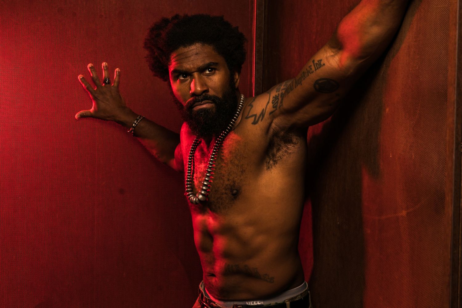 Dramatic portrait in red lighting shows a shirtless person with tattoos posing against a dark red background.